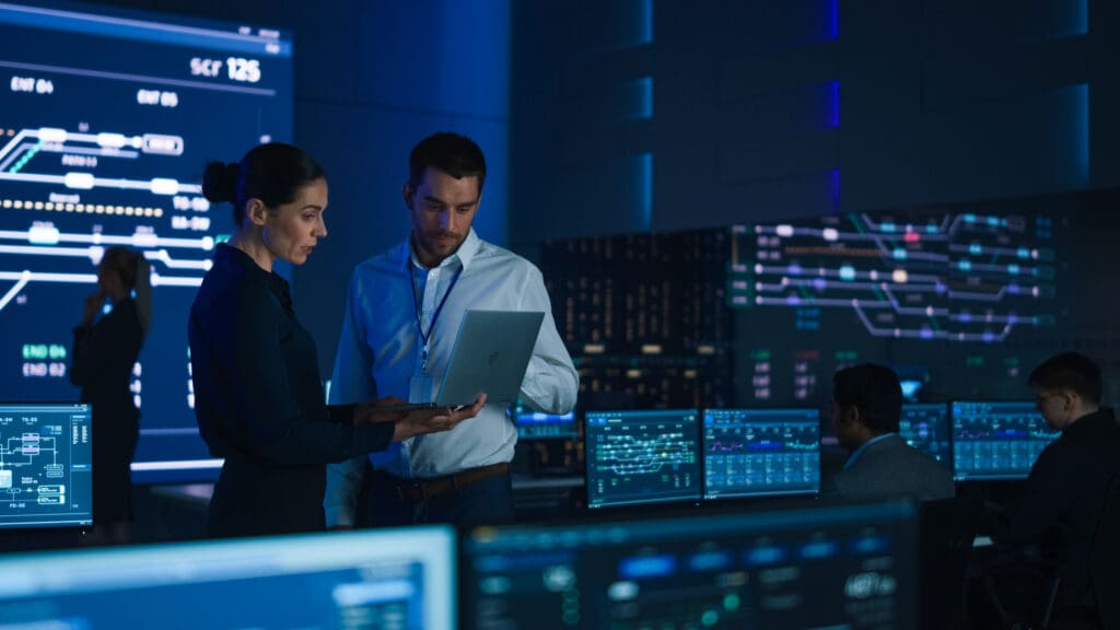 Project leader and boss discuss industrial data on a laptop, with large screen in background.