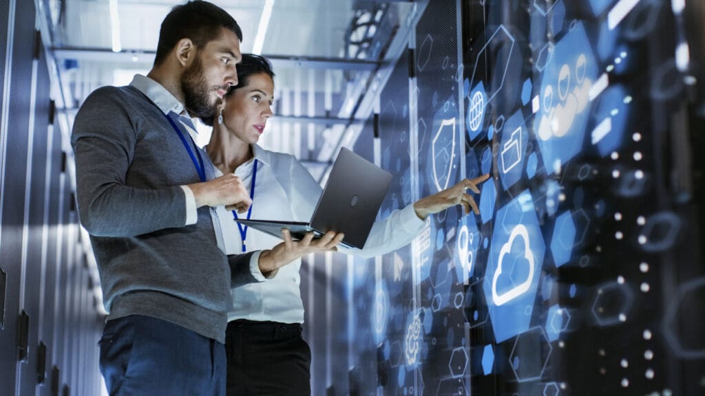 Male IT Specialist Holds Laptop and Discusses Work with Female Server Technician. They're Standing in Data Center, Rack Server Cabinet with Cloud Server Icon and Visualization. Managed PKI solutions mitigate risks